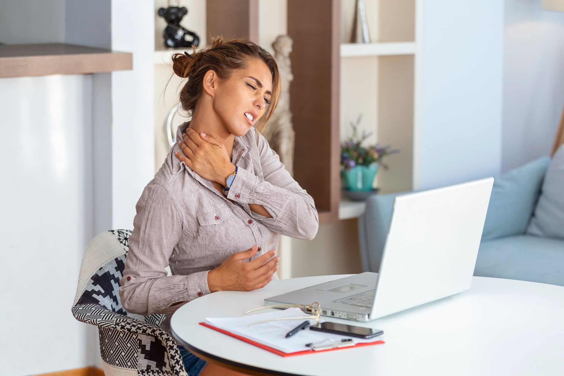A woman is sitting at a desk with a laptop and holding her neck