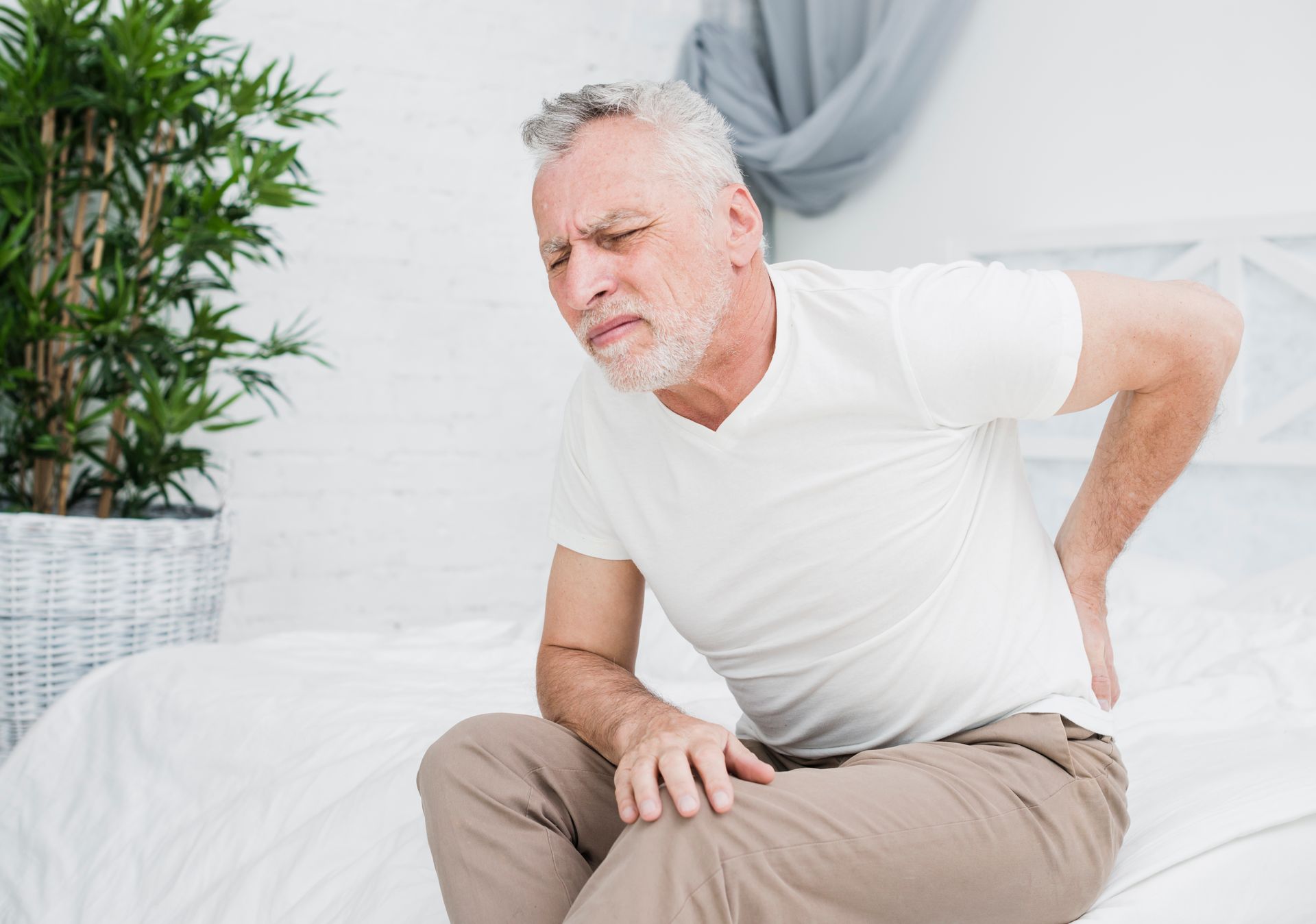 An older man is sitting on a bed holding his back in pain