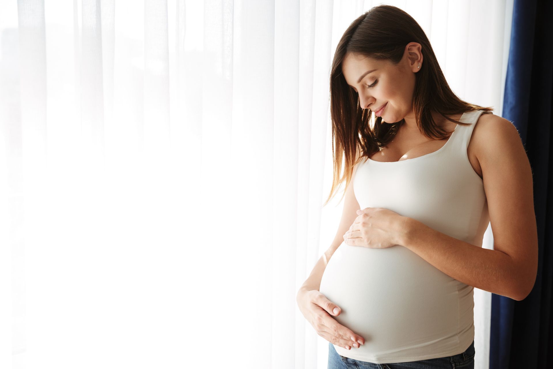 A pregnant woman in a white tank top holds her belly