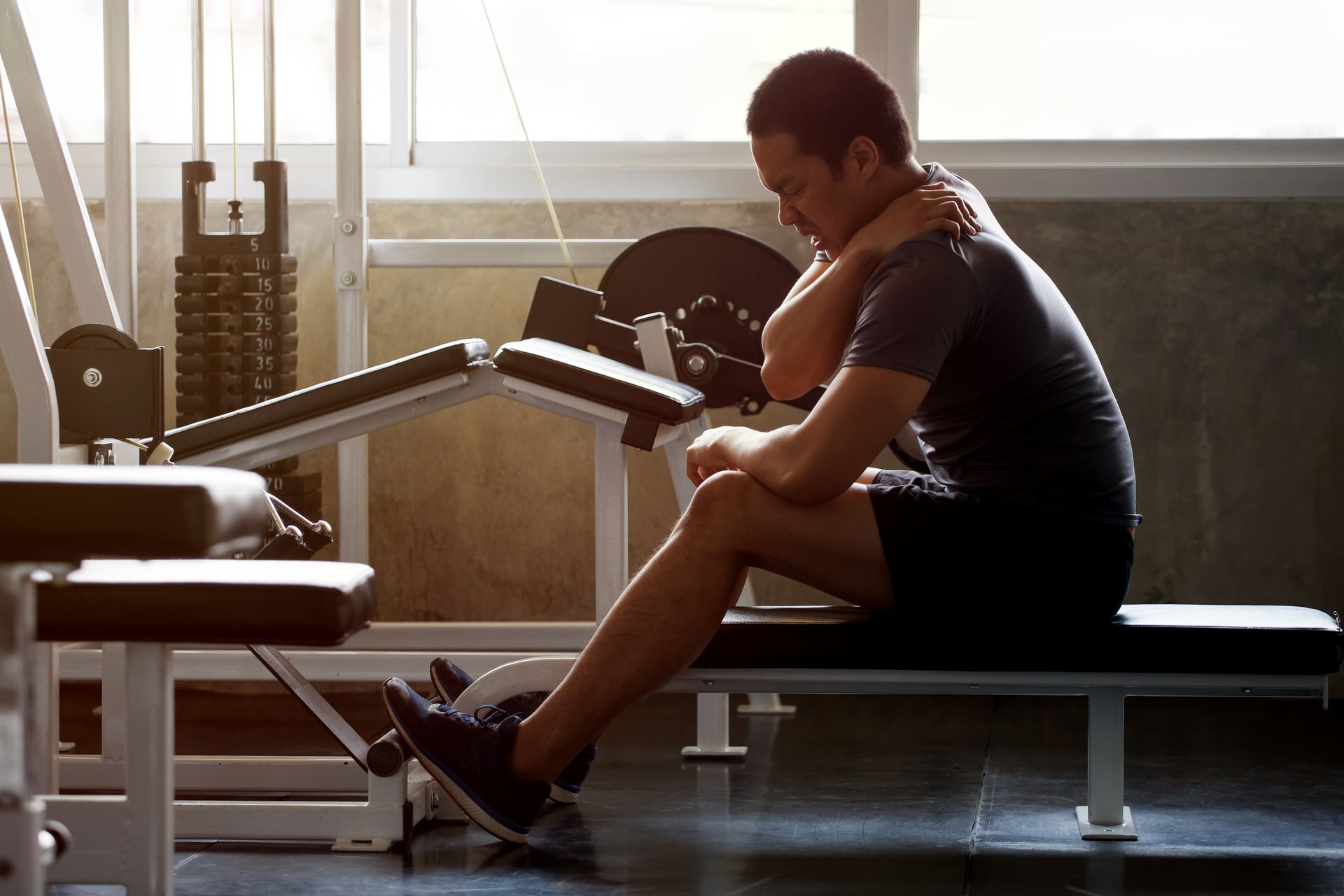 A man sits on a bench in a gym holding his neck
